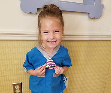 Braylee, a young girl, about 4-5, with dirty blonde hair wearing scrubs smiles into the camera and shows off a craft that she’s made